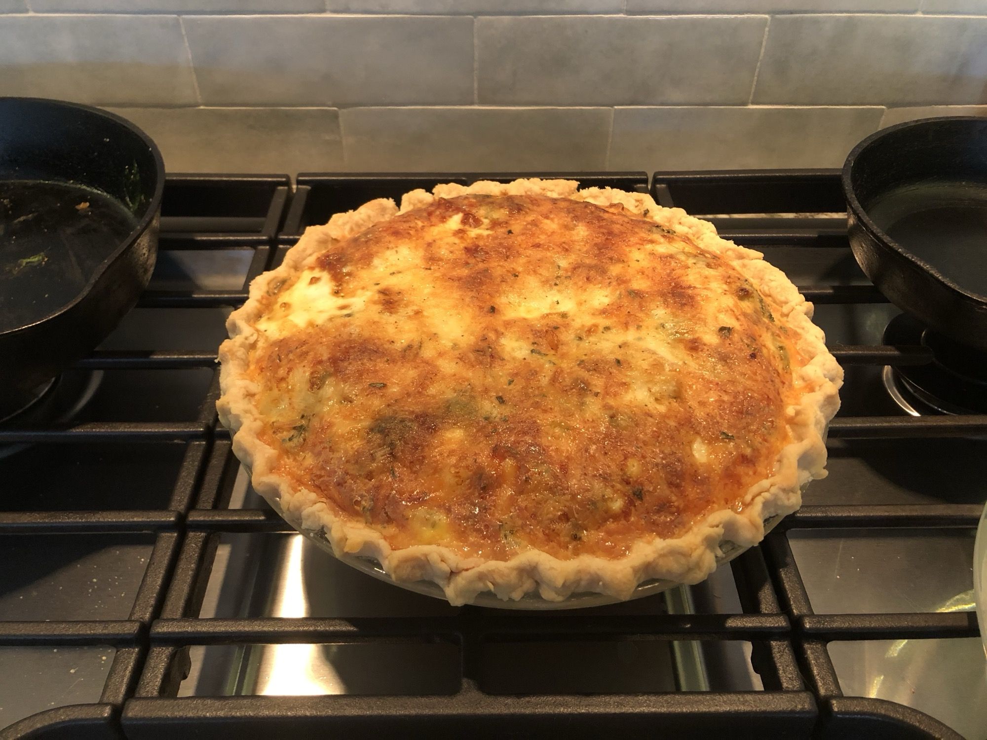 A deep golden-brown quiche with a browned cheese-and-herb custard top in a fluted glass pie plate, cooling on a stovetop.