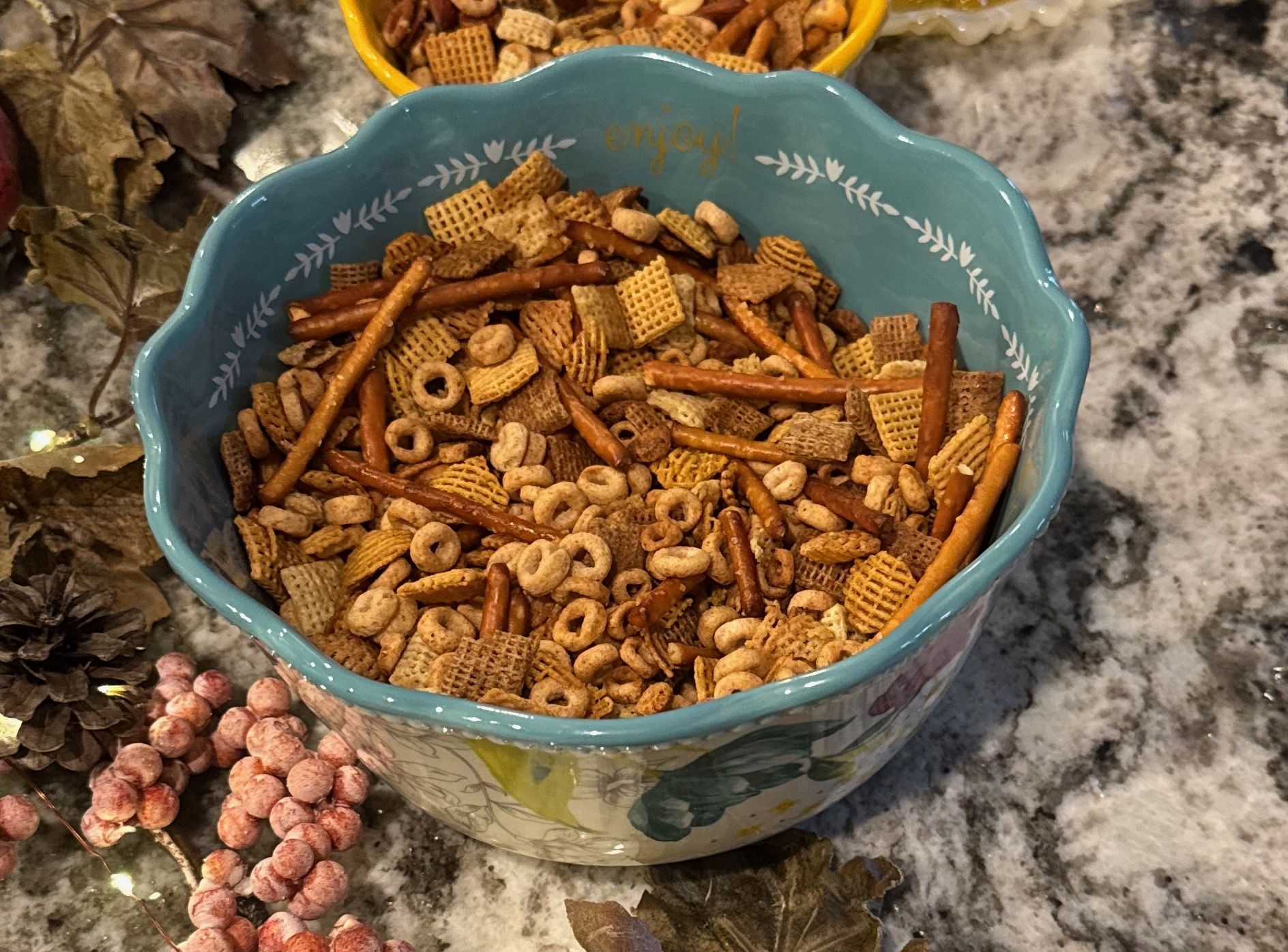 A baked Chex-and-Cheerios snack mix with pretzel sticks in a scalloped ceramic bowl, surrounded by autumn leaves and frosted berries.