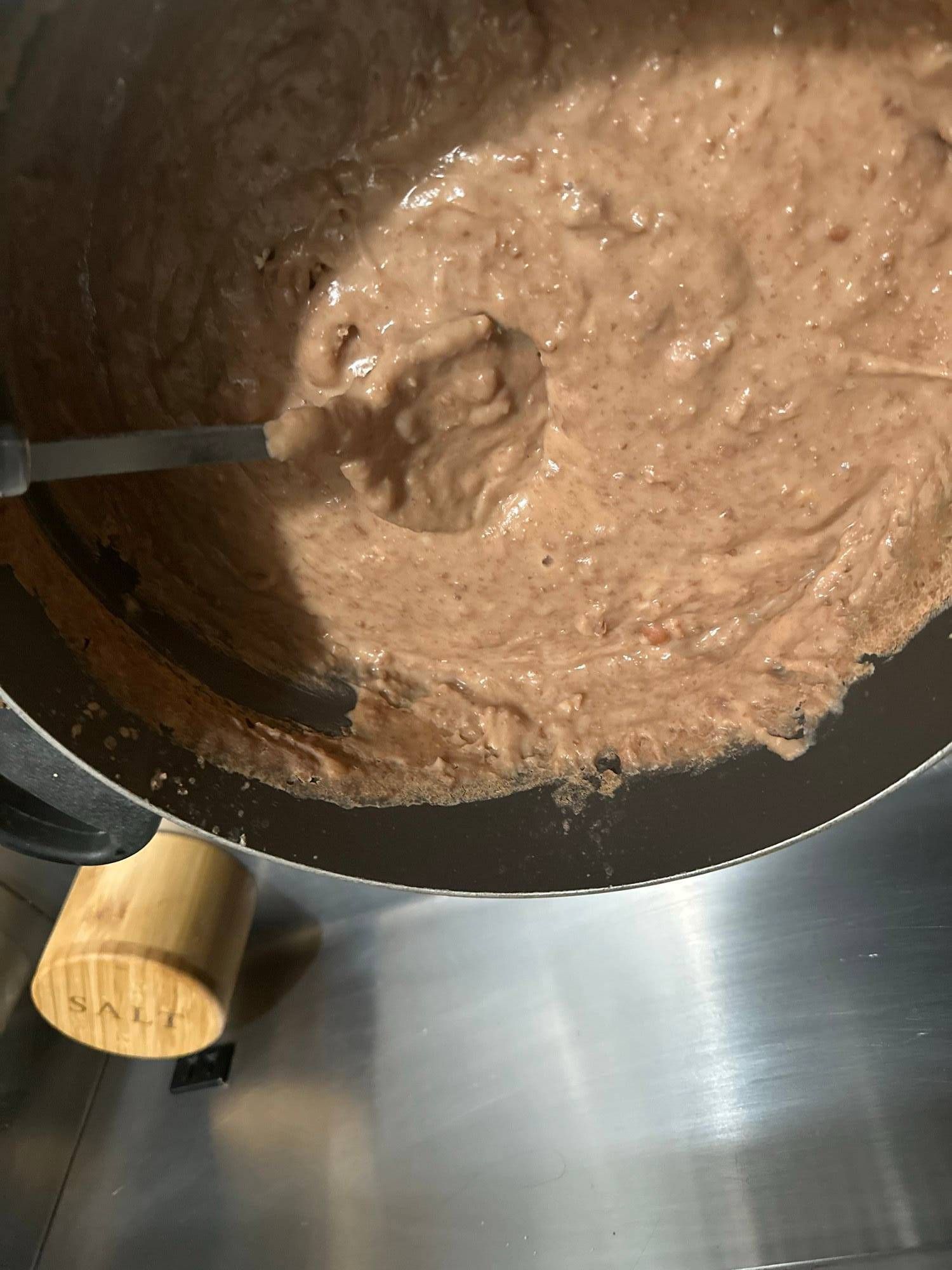 A large pot of creamy, freshly blended refried beans being stirred with a ladle on a stainless-steel counter.