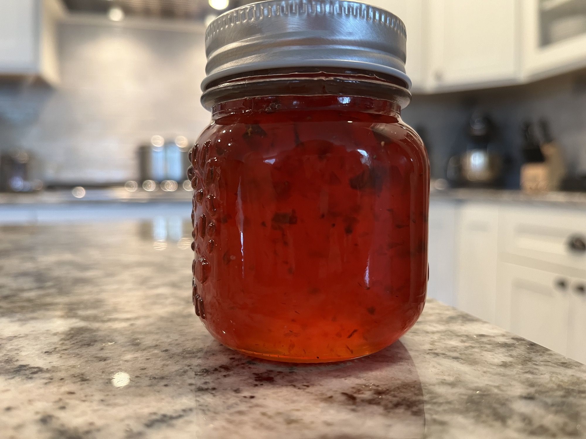 A jar of translucent red-amber jalapeño pepper jelly flecked with peppers, sealed with a canning lid on a granite countertop.